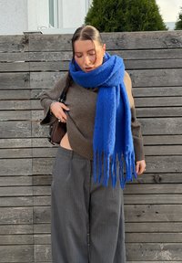 Brun strikket sweater med opkørte ærmekanter, mørkegrå bukser med tynde striber og et lyseblåt tørklæde med frynser. Udendørs træbaggrund.