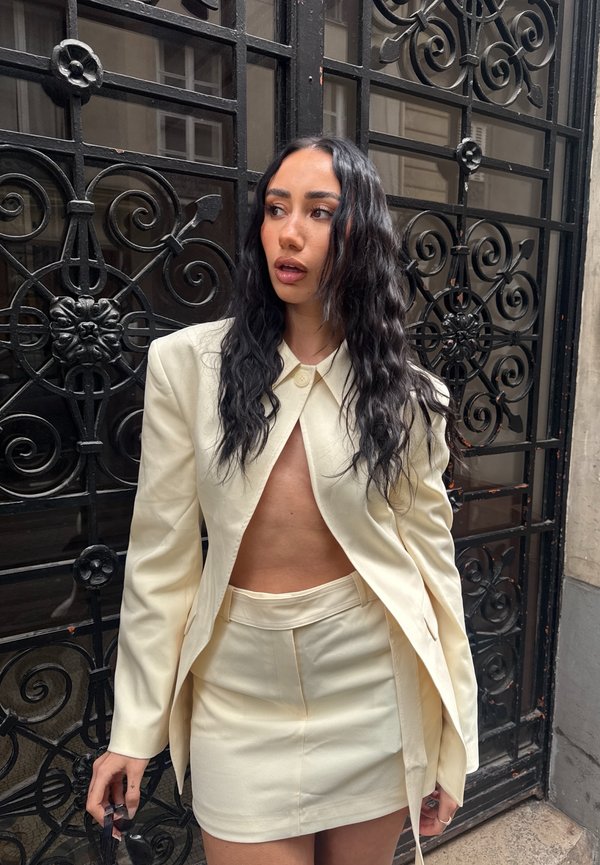 Femme aux longs cheveux ondulés et foncés, portant un blazer crème ouvert et une jupe assortie, se tient devant un portail métallique décoratif noir en regardant sur le côté.