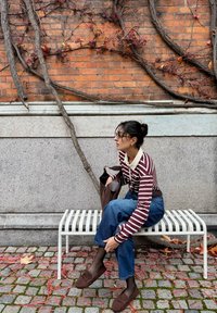 Gestreept shirt met lange mouwen en een beige kraag, blauwe spijkerbroek, bruine schoenen, zittend op een witte metalen bank tegen een bakstenen muur met klimop.