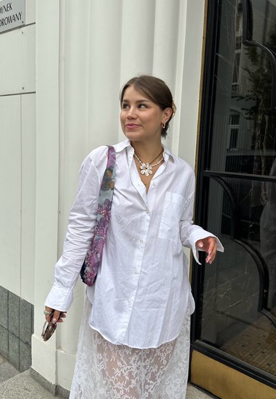 Camisa blanca de gran tamaño con botones, combinada con una falda de encaje transparente, acentuada con un collar floral y un bolso de estampado colorido.