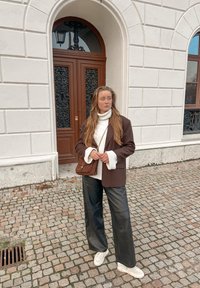 Blazer marron sur un col roulé blanc, pantalon large en cuir noir, et baskets blanches. Sac marron et mur en pierre texturée en arrière-plan.