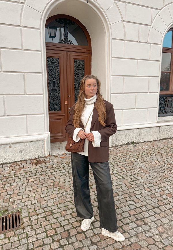 Brun blazer over hvit turtleneck, vide svarte skinnbukser, og hvite joggesko. Brun veske og teksturert steinvegg i bakgrunnen.