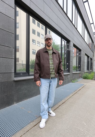 Hombre con chaqueta marrón, camisa verde a rayas, vaqueros azul claro, zapatillas blancas y gorra gris, de pie en la acera junto a un edificio moderno con grandes ventanales.