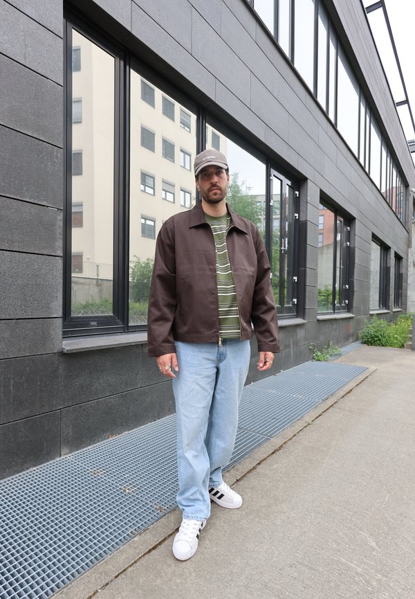 Mann trägt braune Jacke, grün gestreiftes Hemd, hellblaue Jeans, weiße Turnschuhe und graue Kappe, steht am Gehsteig bei einem modernen Gebäude mit großen Fenstern.