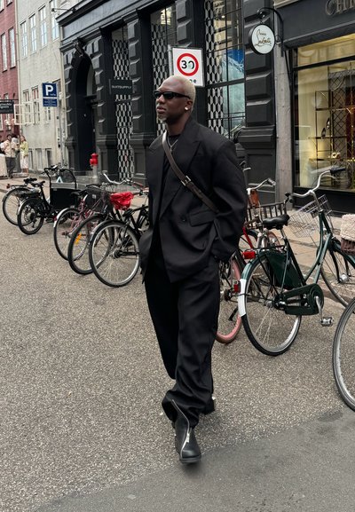 Traje negro a medida con pantalones anchos y una chaqueta oversize. Combinado con botas negras con cremallera y gafas de sol, complementado con un bolso de hombro.