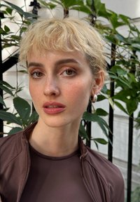 Braune Zip-Jacke über einer taillierten braunen Bluse, mit kurzen blonden Haaren und Creolen, vor grünem Laub und einem schwarzen Tor.