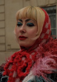 Czerwona, dziergana szal z wyraźnym wzorem w kratę, nałożona na puszystą, wielokolorową tkaninę. Charakteryzuje się intensywnym niebieskim makijażem oczu i wieloma kolczykami na twarzy.