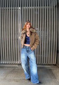 Jeune femme aux cheveux roux portant une veste à col en fourrure marron, un haut bleu à paillettes, un jean à jambes larges et des chaussures noires, debout devant un fond de barres métalliques.