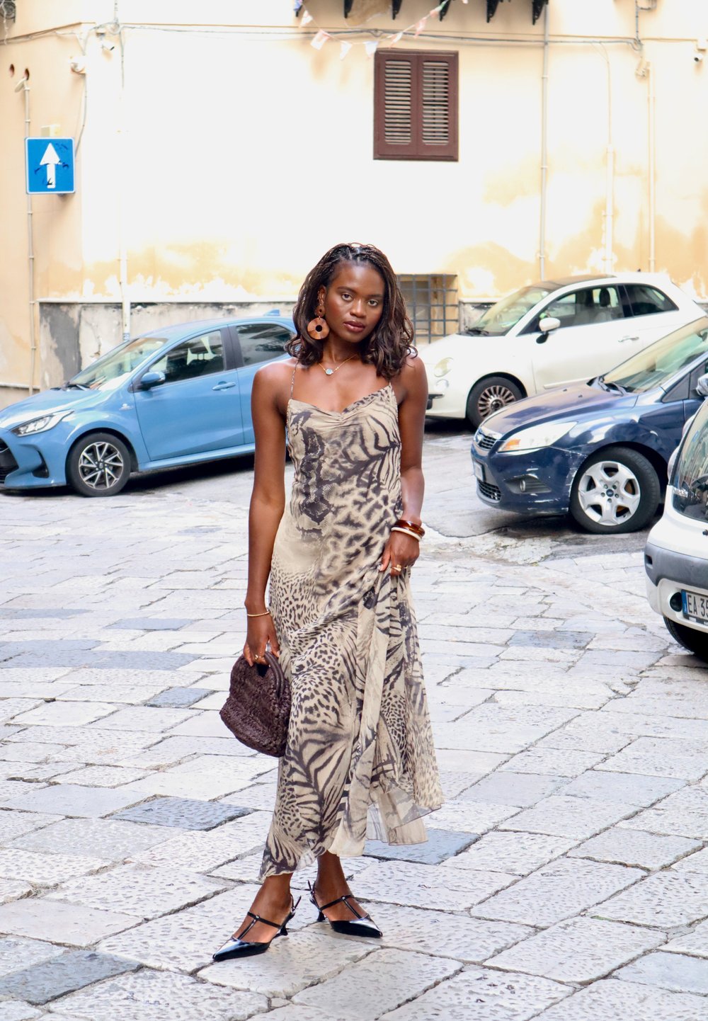 Beige- ja pruunikas loomamustriga voogav kleit, õhukeste rihmadega. Aksessuaaridena punutud pruun kott ja mustad teravnurkadega madalad kingad. Linnakeskkond taustal.