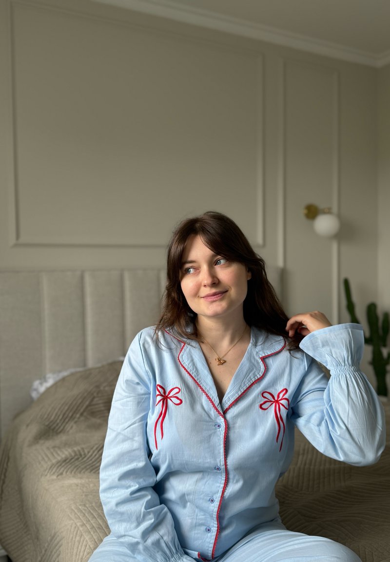 Jeune femme en pyjama bleu avec des nœuds rouges, assise sur un lit dans une chambre doucement éclairée, touchant délicatement ses cheveux et regardant sur le côté.