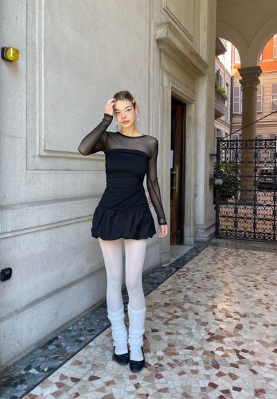 Top negro de malla de manga larga con un corsé ajustado y un dobladillo con volantes, combinado con medias blancas y calcetines de rayas blancos hasta la rodilla. Zapatos negros.