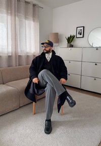 Homme portant des lunettes, une casquette, un manteau et une cravate, assis jambes croisées sur une chaise en bois dans un salon moderne avec un canapé, un tapis gris et des armoires blanches.