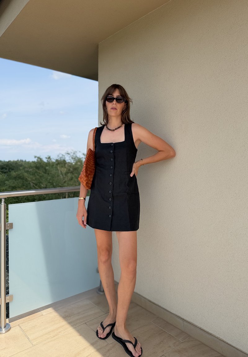 Donna in piedi sul balcone che indossa un vestito nero con bottoni, occhiali da sole neri, infradito, che porta una borsa marrone intrecciata, con alberi e cielo sullo sfondo.