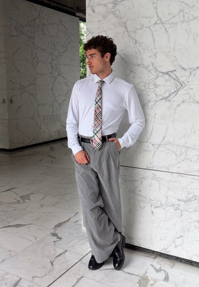 Hombre con camisa blanca, corbata con patrón y pantalones grises con rayas finas apoyado contra una pared de mármol en un edificio moderno con suelo de mármol.