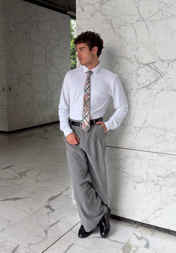 Uomo con camicia bianca, cravatta fantasia e pantaloni grigi a righe sottili che si appoggia a un muro di marmo in un edificio moderno con pavimento in marmo.