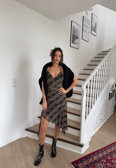 Mujer con un vestido asimétrico de cuadros y botas negras posa junto a una escalera blanca, con un cuadro enmarcado en la pared y una alfombra roja con estampado cerca.