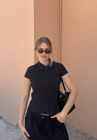 Jovem mulher vestindo blusa preta com fecho, saia preta, óculos de sol retangulares e bolsa preta ao ombro, em pé contra uma parede cor de pêssego.