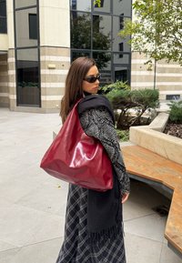 Γυναίκα που φοράει μαύρα γυαλιά ηλίου, μπλούζα με σχέδια και φούστα καρό, κρατώντας μια μεγάλη γυαλιστερή κόκκινη δερμάτινη τσάντα στον ώμο σε αστική αυλή.