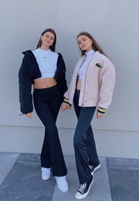 Dwie osoby trzymają się za ręce, ubrane w dopasowane czarne spodnie. Jedna ma na sobie czarną krótką kurtkę, a druga kremową kurtkę sportową z zielonymi akcentami.