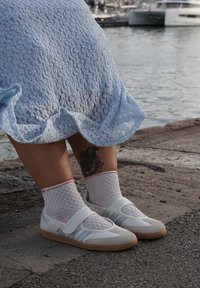 Sapatilhas brancas com detalhes azuis, meias de malha texturizadas com bordas cor-de-rosa, em pé sobre uma superfície de pedra perto de uma massa de água.