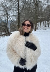 Femme portant un manteau blanc duveteux, des gants noirs et des lunettes de soleil, souriant en plein air dans une forêt enneigée.