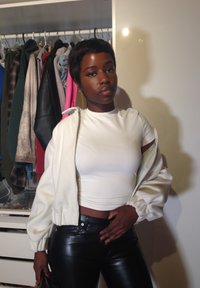 Jonge vrouw in een witte cropped top, witte jas en zwarte leren broek staat voor een open kledingkast met hangende kleren.
