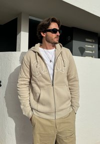 Light beige zip-up hoodie with "GAP" logo, fleece-lined hood, textured fabric, and front pockets, paired with khaki pants.