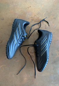 Zwarte sportschoenen met een gestructureerde synthetische bovenkant, voorzien van drie gouden strepen, platte veters en een gevormde rubberen buitenzool voor grip.