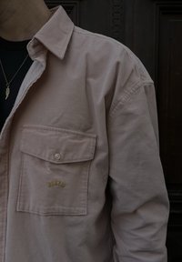 Camisa de bombazina em rosa claro, com corte descontraído, dois bolsos frontais e texto bordado a ouro. Tecido texturizado e design clássico de colarinho.