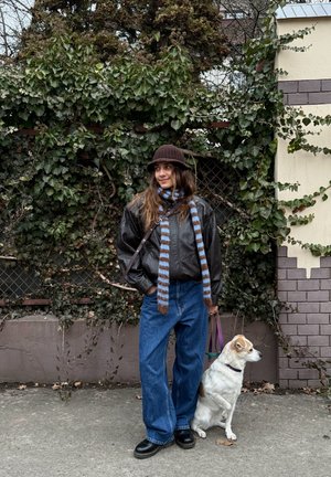 Mujer con sombrero marrón, chaqueta de cuero y bufanda a rayas, de pie en una acera sosteniendo la correa de un perro junto a una pared cubierta de hiedra.