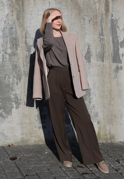 Mujer protegiéndose los ojos del sol, vistiendo un abrigo beige sobre un suéter gris y pantalones anchos oscuros, de pie contra una pared de concreto texturizada.