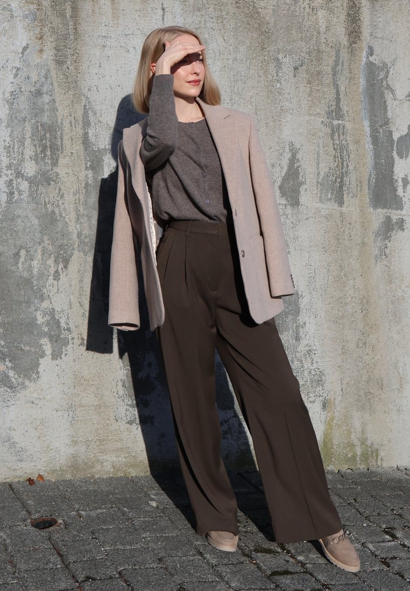 Femme se protégeant les yeux du soleil, portant un manteau beige sur un sweat gris et un pantalon large foncé, debout contre un mur en béton texturé.