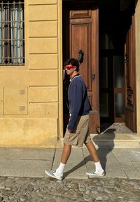 Osoba idąca po brukowanej ulicy, ubrana w granatowy sweter, beżowe szorty, czerwone okulary przeciwsłoneczne i czarno-białe buty wsuwane w kratkę.