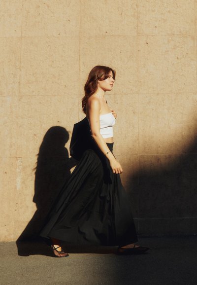 Falda larga negra, top corto blanco y sandalias negras. Tela suave con textura sutil; modelo caminando contra una pared beige, proyectando una sombra.