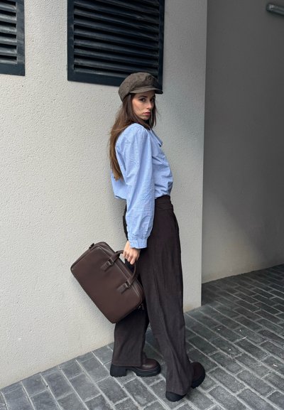 Mujer con blusa azul, pantalones anchos oscuros, botas marrones y sombrero, llevando un maletín marrón, de pie frente a una pared texturizada con rejillas negras.