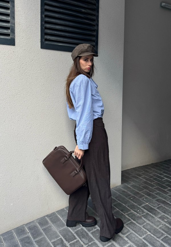 Femme en blouse bleue, pantalon large sombre, bottes marron et chapeau, portant une mallette marron, debout contre un mur texturé avec des grilles noires.