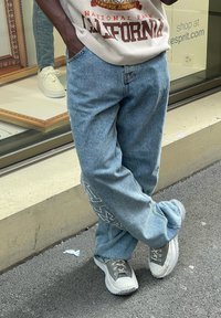 Laza világoskék farmernadrág fehér hímzett részletekkel, szürke és fehér sneakers-szel párosítva. A háttérben egy bolt kirakata tükörrel.