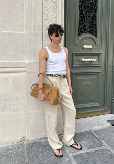 Top blanco sin mangas, pantalones sueltos de color beige, chanclas negras y un bolso marrón texturizado con un detalle de nudo, frente a una puerta histórica.