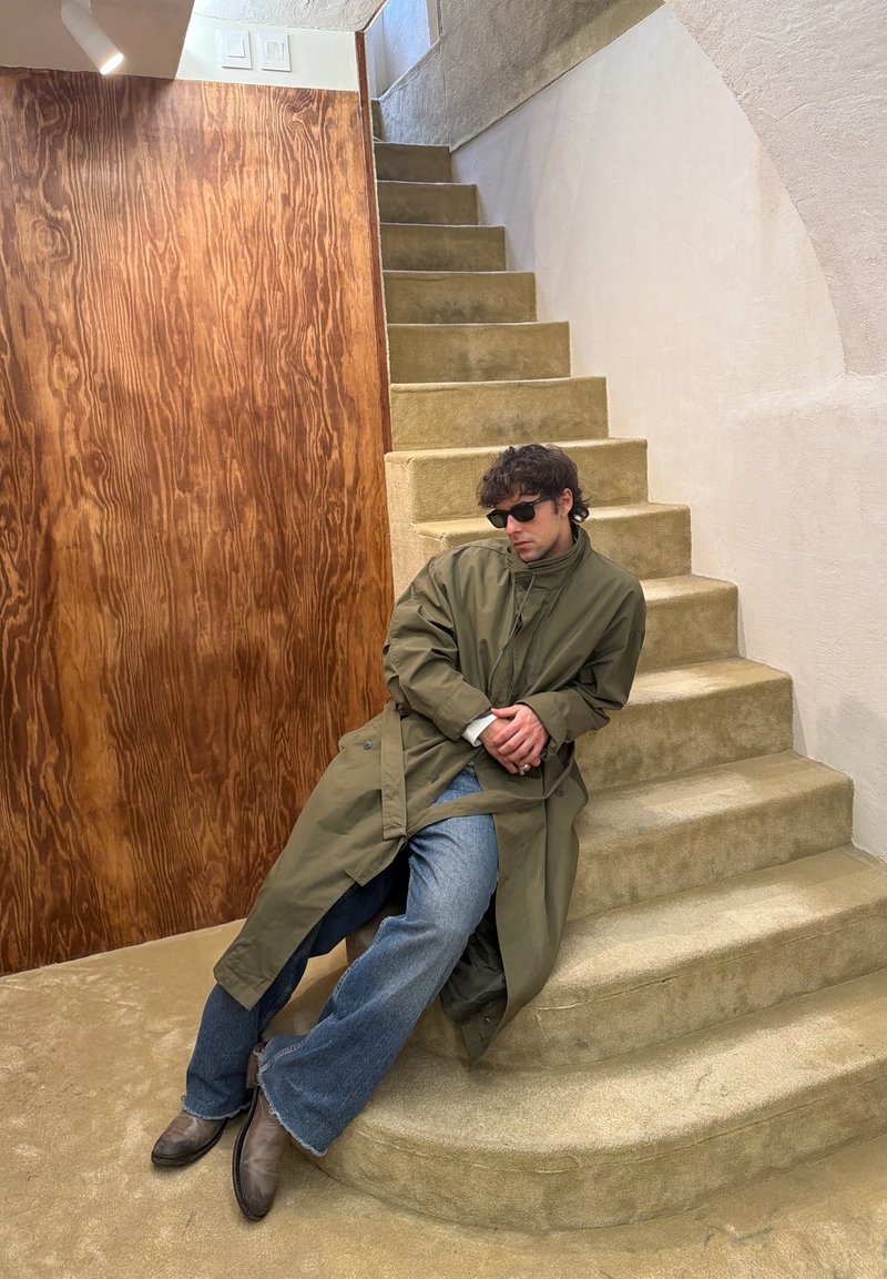 Homme portant des lunettes de soleil, un trench-coat vert, un jean et des bottes, s'appuyant sur un escalier recouvert de moquette beige à côté d'un mur à texture bois.