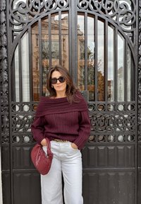 Bordó žebrovaný svetr s odhalenými rameny, nošený s bílými kalhotami širokých nohavic a krčenou bordó taškou, na pozadí zdobené černé brány.