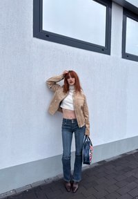 Light beige suede jacket with embossed text, white cropped turtleneck, dark denim flared jeans, and brown pointed-toe boots. Handbag featuring a flag design.