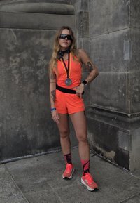 Svijetlo narančasta sportska odjeća s crnim akcentima, s medaljom koja visi s vrpce oko vrata; stoji protiv teksturirane kamenite pozadine.