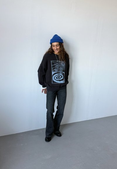 Sudadera negra con diseño gráfico azul, de ajuste holgado. Jeans acampanados gris oscuro. Gorro de punto azul. Botas negras. Fondo sencillo.