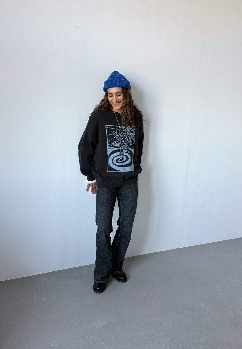 Sweat-shirt noir avec un motif graphique bleu, coupe ample. Jean évasé gris foncé. Bonnet tricoté bleu. Bottes noires. Fond simple.