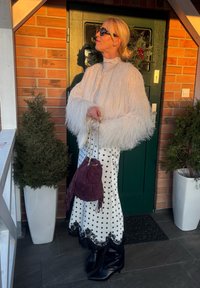 Femme portant une veste blanche duveteuse, jupe à pois avec dentelle noire, bottes noires, lunettes de soleil, tenant un sac à main violet, debout près d'une porte verte avec une couronne.