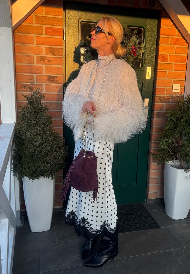 Femme portant une veste blanche duveteuse, jupe à pois avec dentelle noire, bottes noires, lunettes de soleil, tenant un sac à main violet, debout près d'une porte verte avec une couronne.