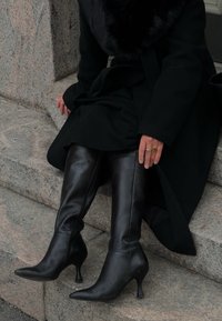 Térdig érő fekete bőr csizma hegyes orral és vékony sarkakkal. Tiszta textúra és minimalista design, fekete kabáttal párosítva.