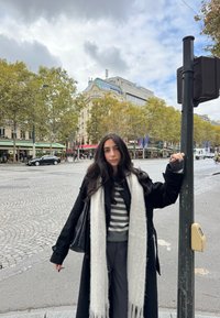 Črni plašč z velikimi gumbi, sivi pulover z vzorcem črt, svetlosiva ruta in temne hlače. Ulični tlak iz tlakovcev in mestna kulisa z drevesi.