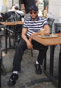 Camisa polo a rayas en azul marino y blanco, pantalones negros, gafas de sol redondas y zapatos negros con suela gruesa, sentado en una mesa de madera con un café.