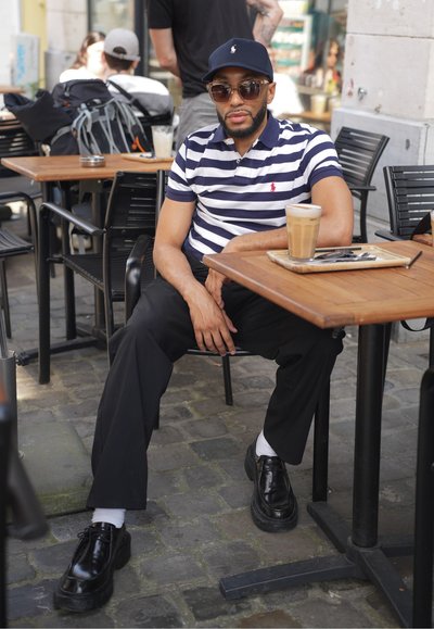 Camisa polo a rayas en azul marino y blanco, pantalones negros, gafas de sol redondas y zapatos negros con suela gruesa, sentado en una mesa de madera con un café.